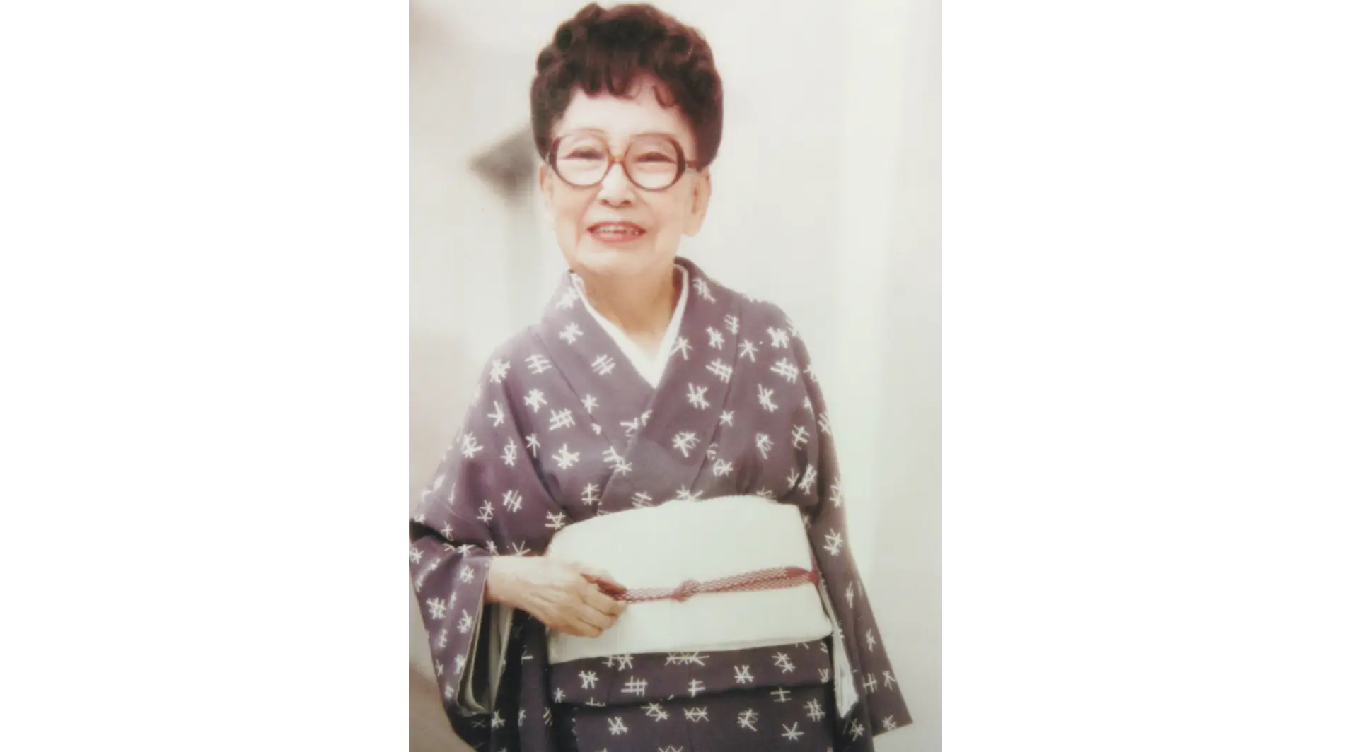 井桁(いげた)模様の着物に白い帯を締め、丸眼鏡をかけて優しくカメラに微笑みかける宇野千代のポートレート写真