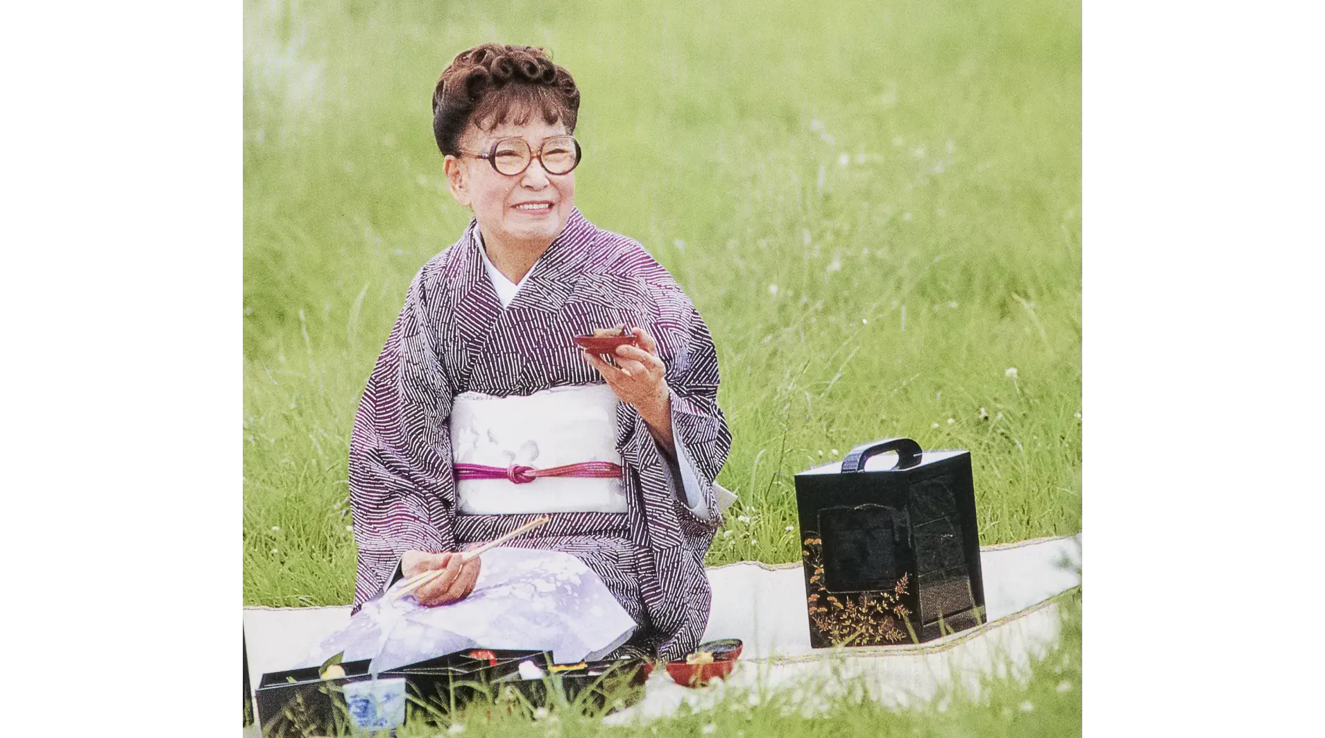 青々とした芝生の上でシートを敷き、提げ重箱やお弁当を広げて盃を手に野外での食事を楽しむ宇野千代