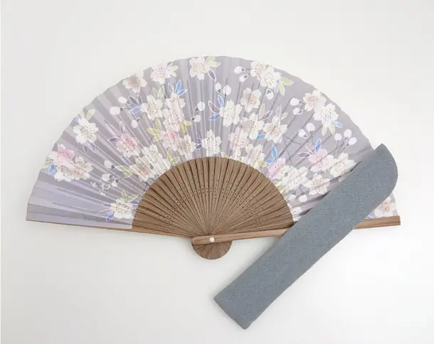 淡いグレーの地に白やピンク、紫の桜の花々が繊細に描かれた上品なデザインの扇子。右下には扇子と同色の無地の扇子袋が斜めに添えられている