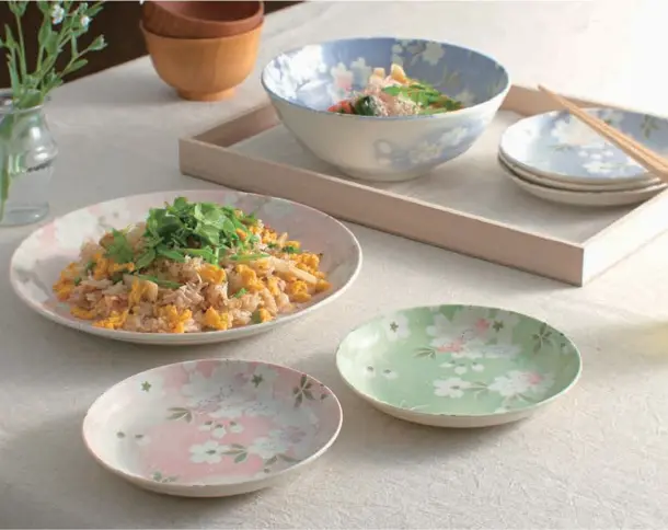 食卓に並ぶ宇野千代デザインの食器類。手前にはピンクとグリーンの淡い桜柄の小皿、奥にはピラフが盛られた大皿、木製トレイの上にはスープが入ったブルーのボウルや箸が添えられたお皿が並ぶ