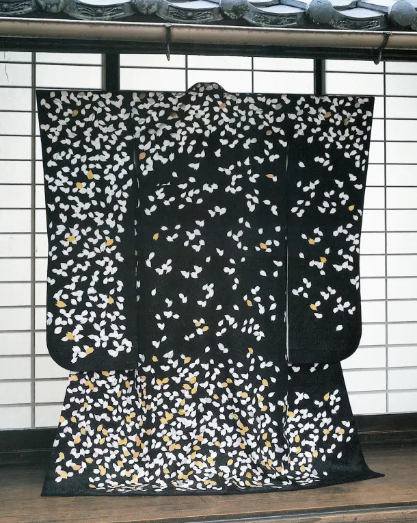 伝統的な日本家屋の瓦屋根と白い障子の前に美しく広げて掛けられた、漆黒のちりめん地に無数の白い桜の花びらがリズミカルに舞い散るシックな和の情緒漂う宇野千代デザインのきもの