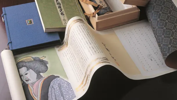 手前に広げられた『おはん』の浮世絵風挿絵や直筆原稿が印刷された美しい巻物を中心に、奥には『大狗庵久吉 宇野千代』と書かれた緑色の書籍、青色の特製本、木箱などの貴重な文学資料が並ぶ画像