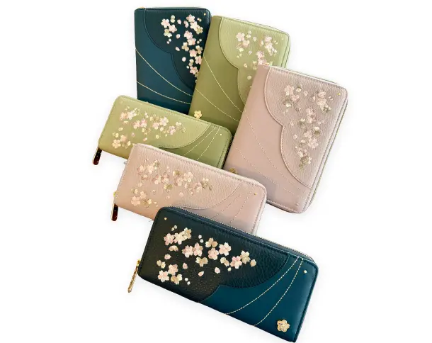 上品なシボ革に桜の花々がゴールドやピンクの箔押しと美しい立体刺繍で描かれた、L字またはラウンドファスナー式の長財布と折り財布。深緑、若草色、くすみピンクの3色のバリエーションが並んでいる様子