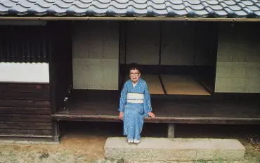 落ち着いた青地に星のような模様が散りばめられた着物を着て、伝統的な日本家屋の瓦屋根がある縁側に腰掛け、優しく微笑む晩年の宇野千代のポートレート写真