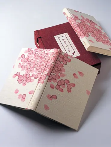 淡いベージュの布地に可憐なピンクの桜の花や花びらが一面に描かれた書籍『生きて行く私』の、美しい装丁本とエンジ色の特製ケース