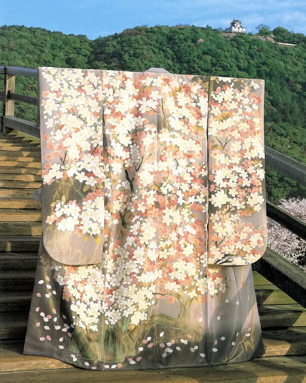 緑豊かな山頂にそびえる城を背景に、歴史ある木製の橋の手すりに美しく掛けられた、上品な薄グレーの生地に白やピンクの桜の花が流れるように満開に描かれた宇野千代デザインの着物