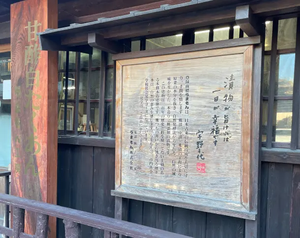 木の風合いを生かした看板。右側には『漬物が旨ければ一日が幸福です 宇野千代』という言葉が書かれた木製プレートがあり、左側には『甘酢白たくあん うまもん工房』と書かれた木製の縦型看板が立てられている