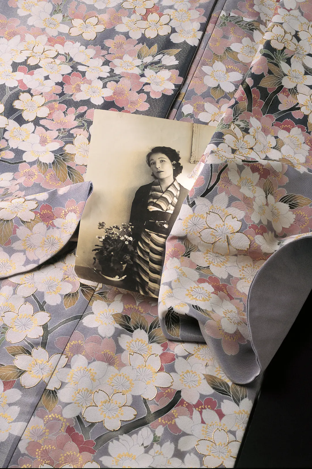 金糸や淡いピンク、白の桜模様が贅沢に織り込まれた華やかな宇野千代デザインの着物の美しく波打つ生地の上に、若い頃の宇野千代のモノクロのポートレート写真が添えられた画像