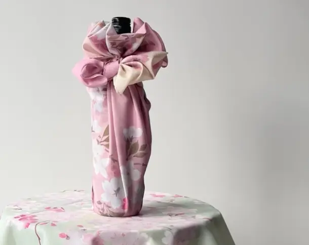 薄緑にピンクの桜が描かれたテーブルクロスの上のボトル。ボトル全体がピンクの桜柄の風呂敷で包まれ、ボトルの首元で華やかなリボン結びに仕上げられている様子